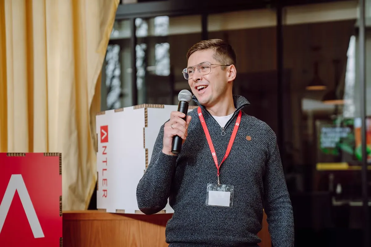 A founder in glasses holding a microphone and smiling at an Antler event, branded signage behind