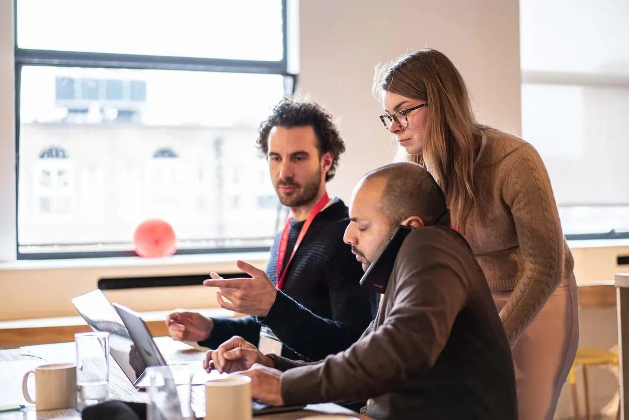 Three founders around a table, one on a phone call while the others review something on a laptop